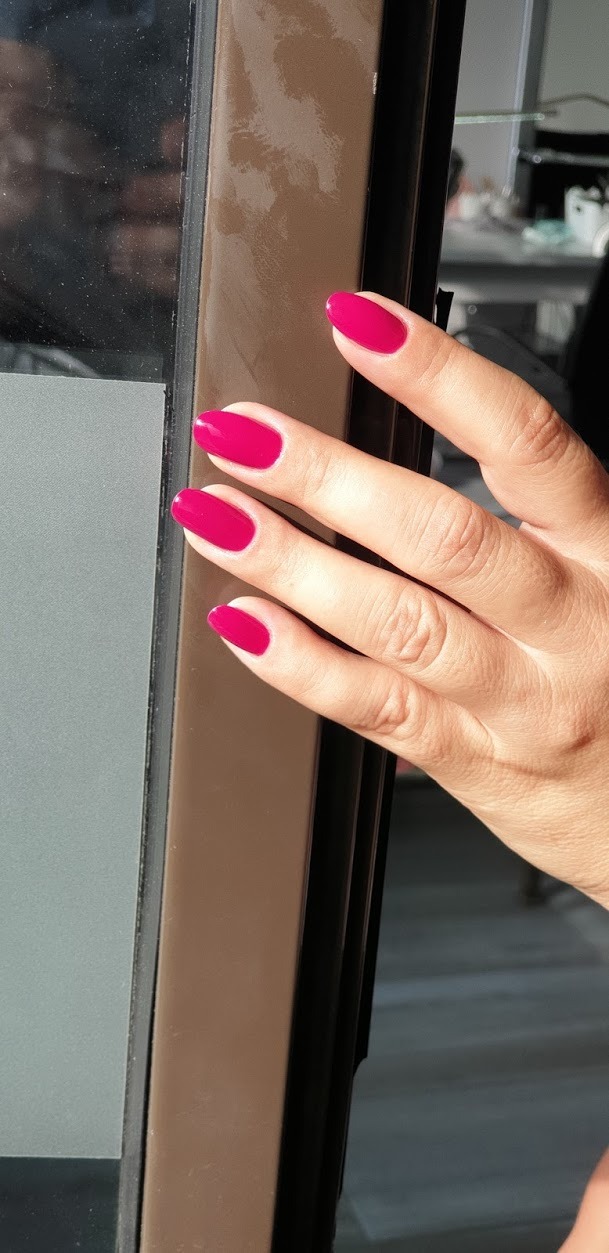 Dłoń z pomalowanymi na intensywny róż paznokciami o owalnym kształcie, na tle ramy okiennej w słoneczny dzień.