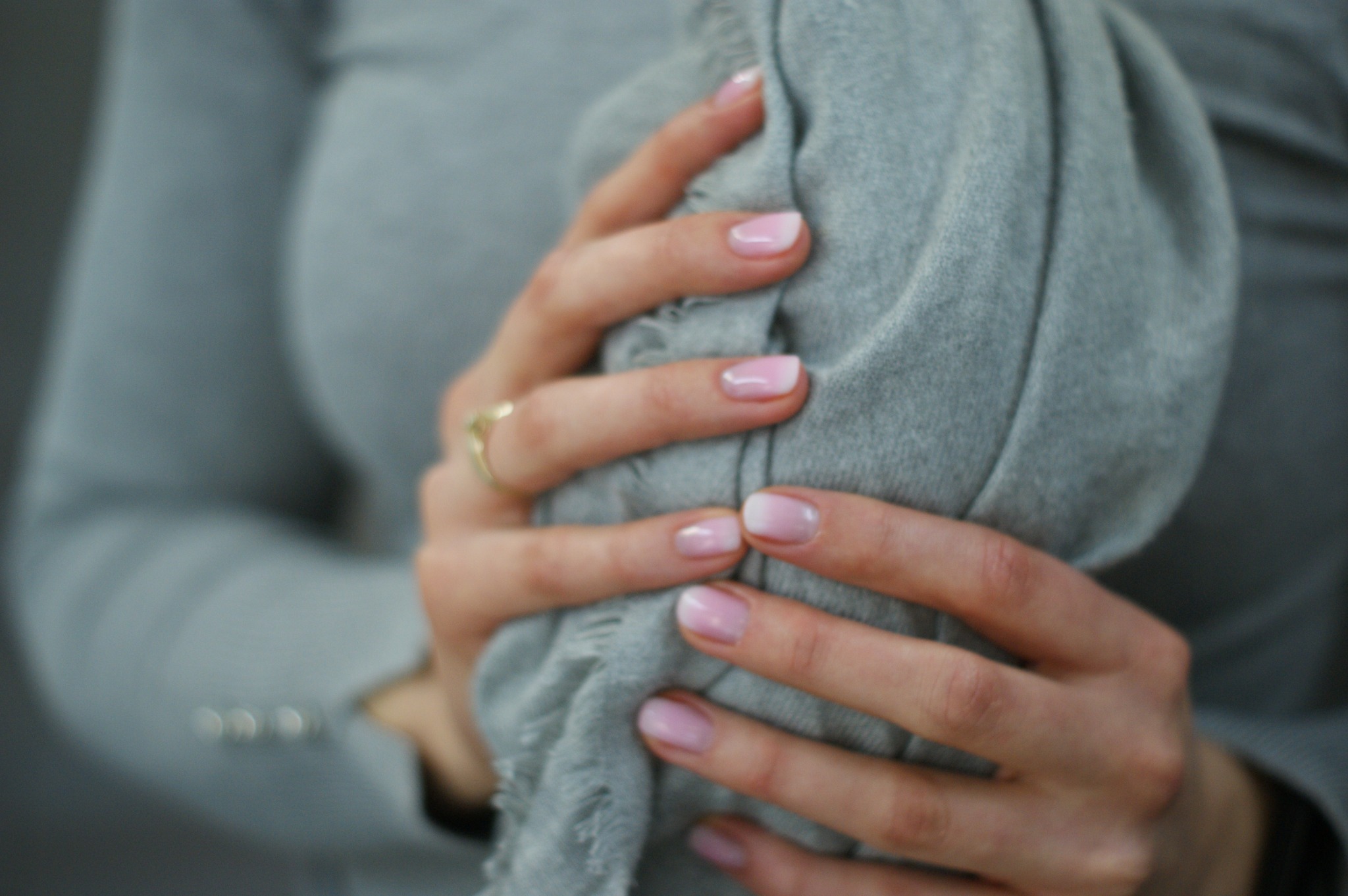 Dłonie kobiety z delikatnym, różowo-białym ombre na paznokciach, trzymające szarą, teksturowaną tkaninę. Na palcu serdecznym złoty pierścionek.