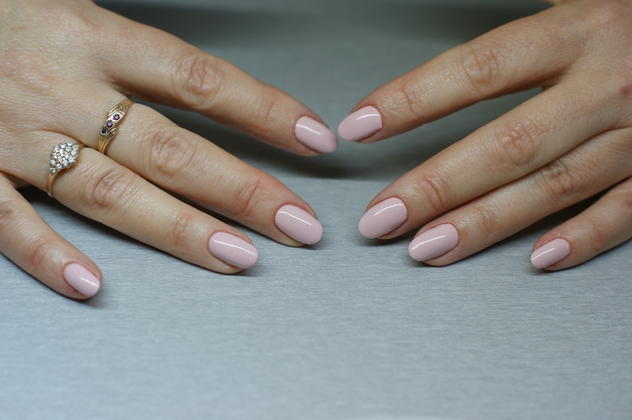 Dłonie z delikatnym, różowym manicure hybrydowym na krótkich, owalnych paznokciach; na palcu serdecznym złoty pierścionek z białymi i czerwonymi kamieniami.