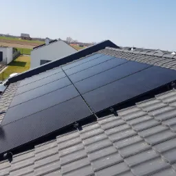 Cztery czarne panele słoneczne zamontowane na szarym dachu z dachówki ceramicznej, widok z góry na tle zabudowy jednorodzinnej i błękitnego nieba.