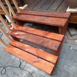 Drewniane schody zewnętrzne z trzema stopniami, świeżo polakierowane na brązowo, zamocowane na śruby, prowadzące do drewnianego podestu tarasu.