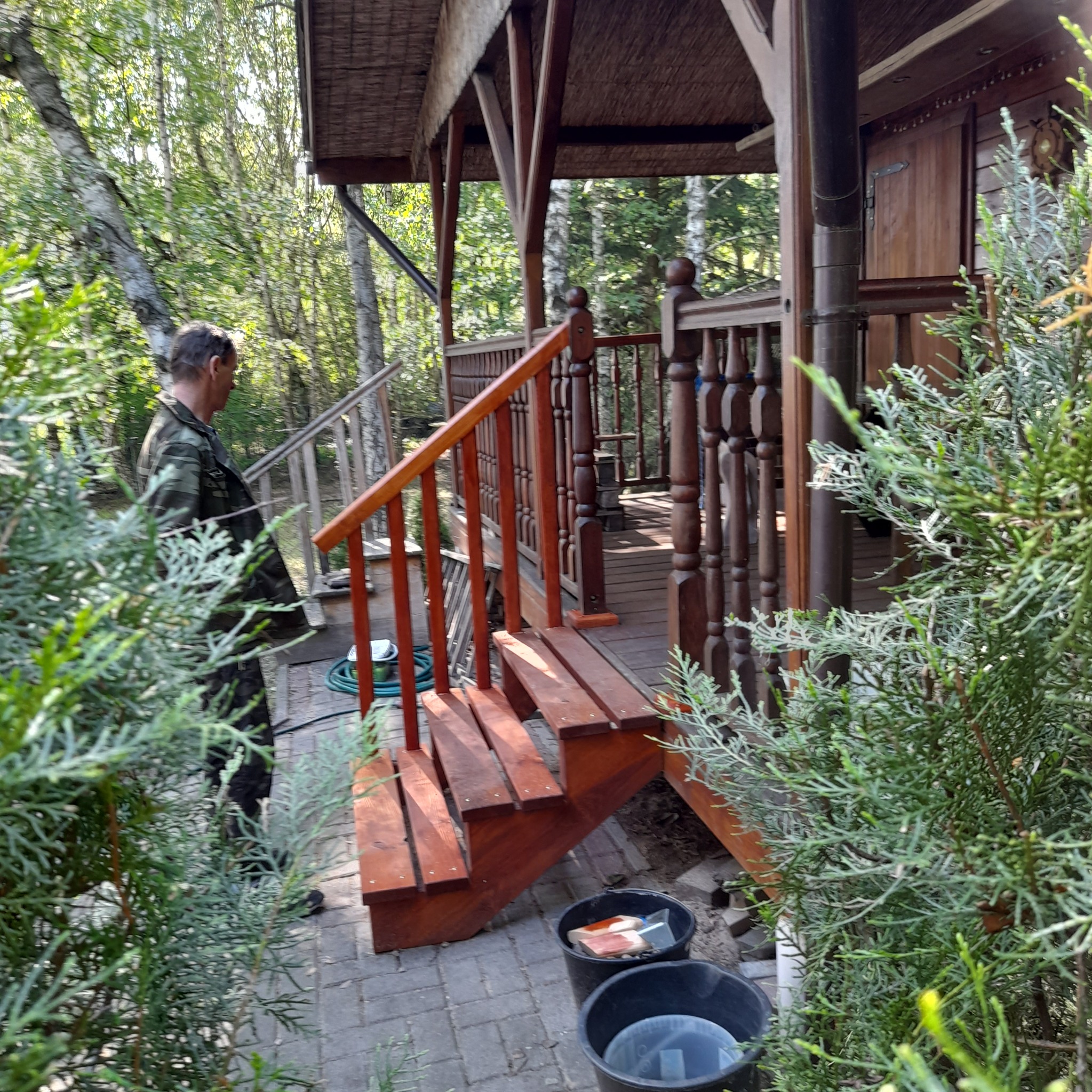 Drewniane schody prowadzące do altany, widoczne dwa wiadra z narzędziami i pozostałościami po malowaniu, mężczyzna w ubraniu roboczym na schodach.