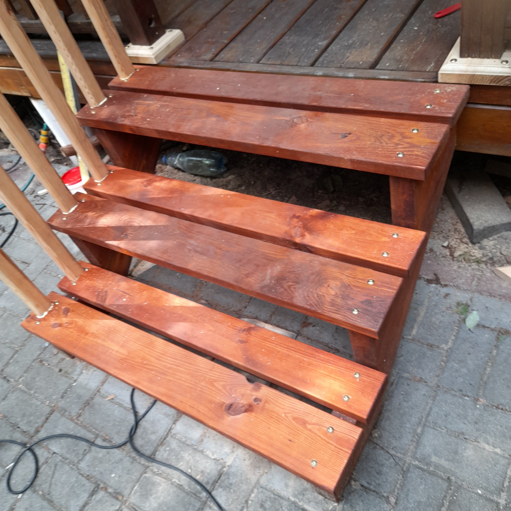 Drewniane schody zewnętrzne z trzema stopniami, świeżo polakierowane na brązowo, zamocowane na śruby, prowadzące do drewnianego podestu tarasu.