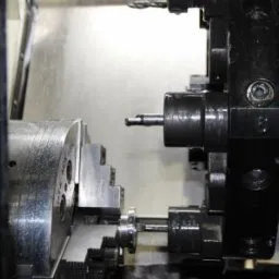 Metalowy detal obrabiany na precyzyjnej tokarce CNC, widoczne narzędzie skrawające i uchwyt mocujący, oświetlenie halogenowe.