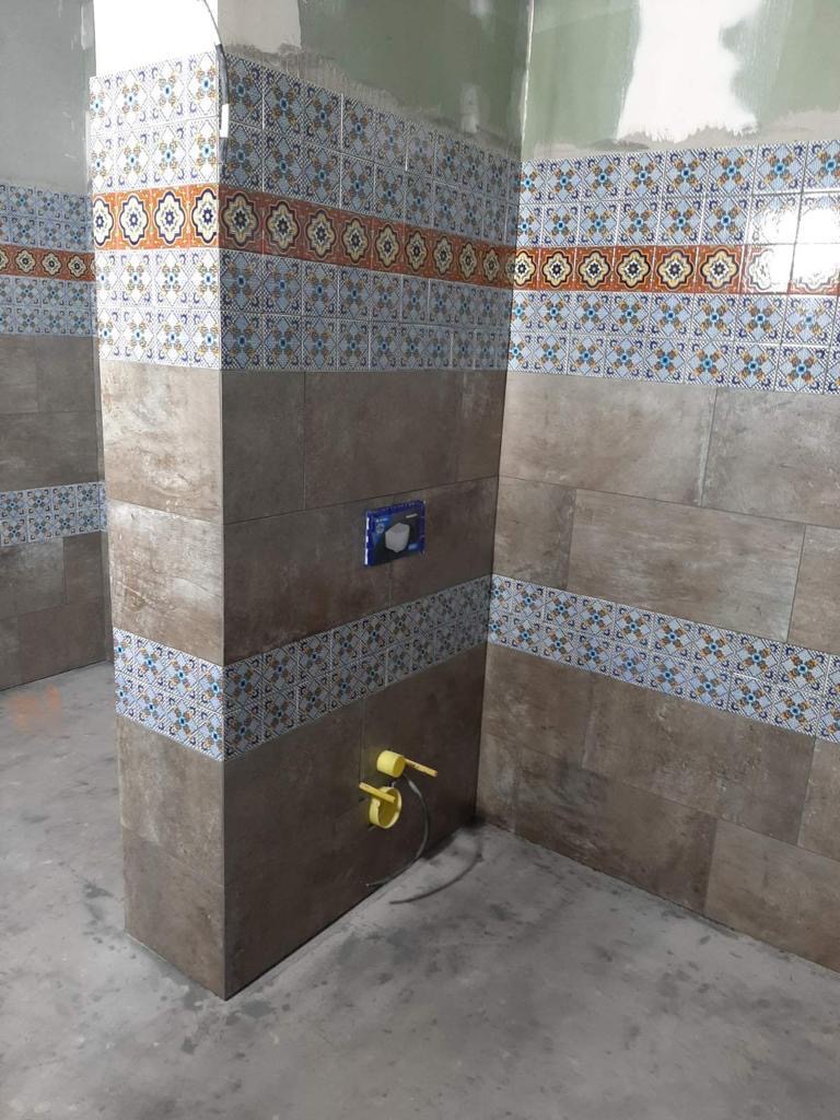 Fragment łazienki w trakcie prac wykończeniowych, z brązowymi płytkami imitującymi drewno oraz dekoracyjnymi pasami płytek z wzorem geometrycznym w odcieniach niebieskiego i pomarańczowego.