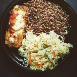 Czarny talerz z porcją: kotlet panierowany z roztopionym serem i sosem pomidorowym, kasza gryczana oraz surówka z kapusty z marchewką.