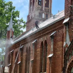 Czyszczenie dachu kościoła z czerwonej cegły pod wysokim ciśnieniem, widoczni dwaj pracownicy w uprzężach, drabina oparta o ścianę, w tle wieża kościelna i drzewa.