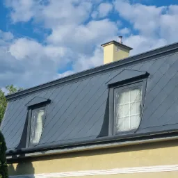 Szary dach mansardowy z dwoma oknami dachowymi zabezpieczonymi folią malarską, widoczny komin i fragment elewacji budynku w słoneczny dzień.