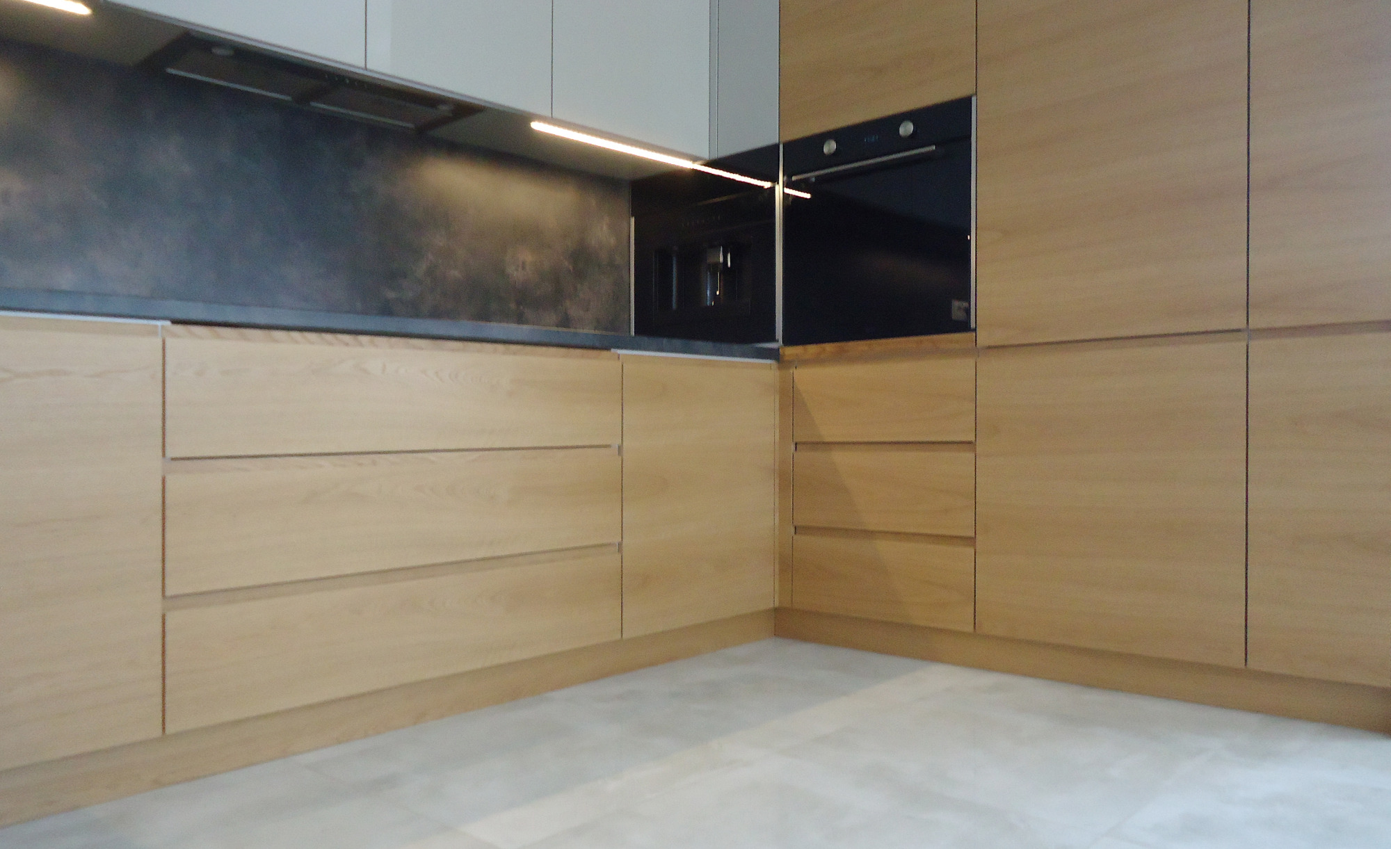 Jasna, drewniana zabudowa kuchenna z czarnym piekarnikiem i ekspresem do kawy, podświetlana listwą LED pod szafkami wiszącymi, na tle szarej ściany.