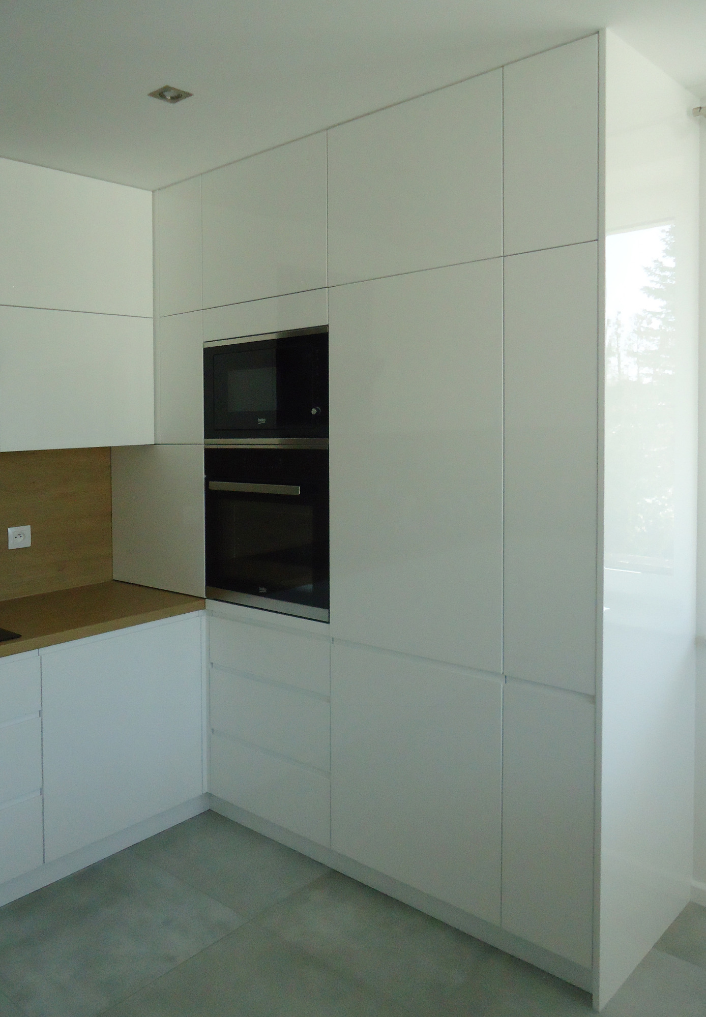 Minimalistyczna, biała kuchnia z zabudową sięgającą sufitu, z wbudowanym piekarnikiem i mikrofalówką, drewniany blat i szare płytki podłogowe.