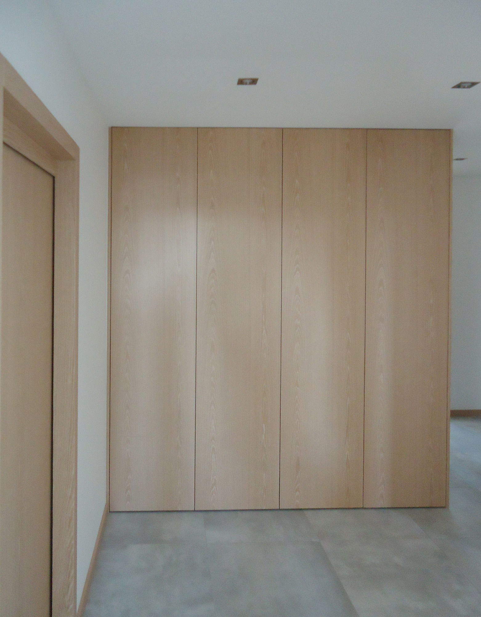 Świetlista, drewniana szafa wnękowa z czterema drzwiami, wkomponowana w ścianę, z subtelnym oświetleniem sufitowym i szarą podłogą, widziana pod kątem z lewej strony pokoju.