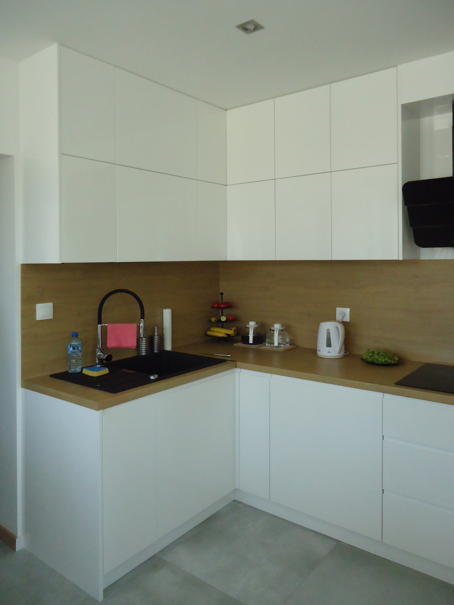 Jasna kuchnia z białymi szafkami, drewnianym blatem i czarnym zlewem z baterią w kształcie łuku, uzupełniona o czajnik i owoce na blacie.