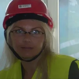 Portret kobiety w okularach, czerwonym kasku z naklejką firmy i odblaskowej kamizelce, na rozmytym tle okna.