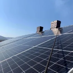 Panele słoneczne na dachu domu z dwoma kominami na tle błękitnego nieba i oddalonych wzgórz.