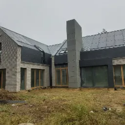 Nowoczesny dom jednorodzinny z szarej cegły i panelami słonecznymi na dachu, widok z zewnątrz na tle zarośniętej działki.