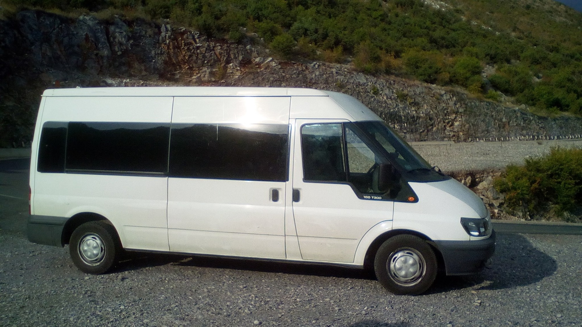 Biały Ford Transit 100 T300 z przyciemnianymi szybami stoi na żwirowym parkingu na tle skalistej góry porośniętej zielenią.