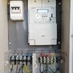 Wnętrze skrzynki elektrycznej z zamontowanym licznikiem energii, bezpiecznikiem i podłączeniem przewodów zasilających. Widoczne zaciski, kable w różnych kolorach i elementy montażowe.