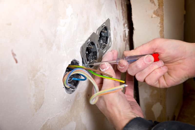 Instalacja gniazdka elektrycznego: dłonie montera sprawdzają napięcie próbnikiem w gniazdku, widoczne kolorowe przewody i surowa ściana w tle.
