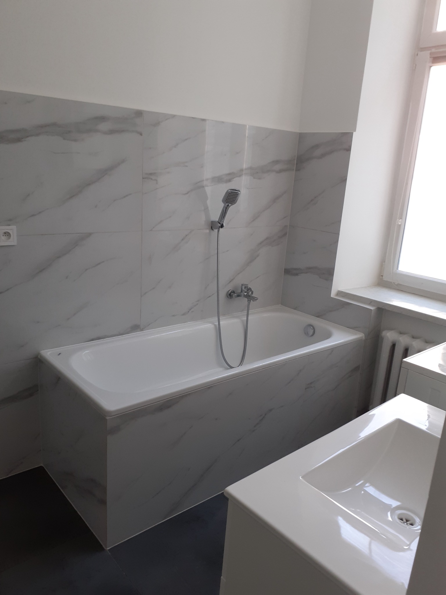 Wyremontowana łazienka z wanną obudowaną płytkami imitującymi marmur, armaturą natryskową i umywalką w zabudowie, widok na fragment okna i grzejnika.