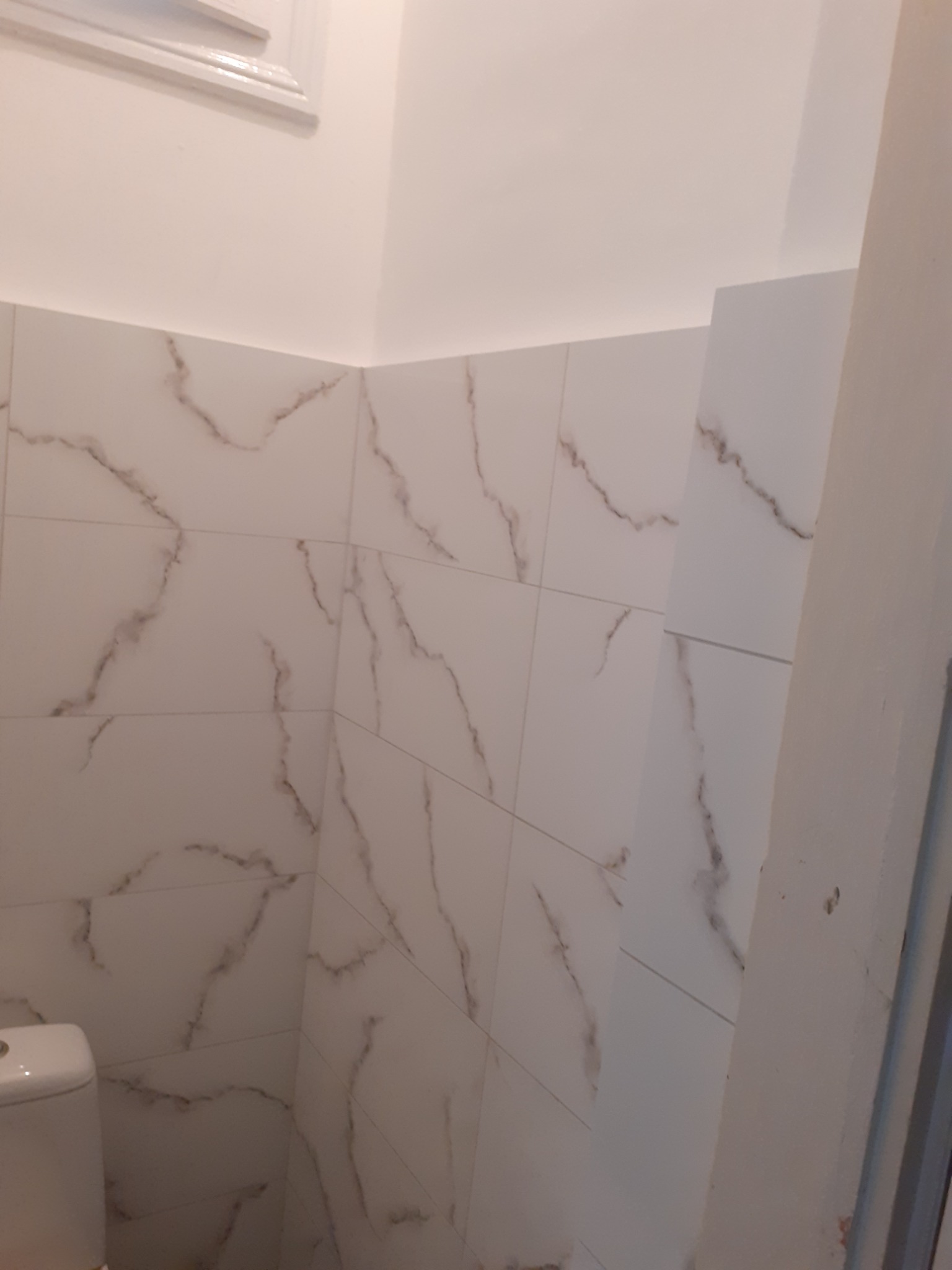 Wykończona ściana w łazience z białymi płytkami imitującymi marmur, widoczny fragment toalety i okna.