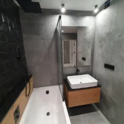 Nowoczesna łazienka z szarą płytką imitującą kamień, czarną armaturą i drewnianymi akcentami, kabina prysznicowa ze szklaną ścianką, widoczne odbicie w lustrze.