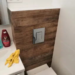 Nowoczesna toaleta z drewnopodobną obudową stelaża, chromowanym przyciskiem spłukującym, białą ceramiką i płytkami, z widocznym fragmentem umywalki oraz butelką szamponu.