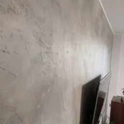 Ściana pokryta dekoracyjną szarą gładzią w stylu betonowym, widoczny fragment telewizora i mebla w tle.