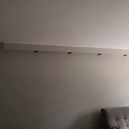 Jasnoszara ściana z zamontowanym pod sufitem podwieszanym elementem z czterema punktowymi oprawami oświetleniowymi, fragment szarej sofy widoczny w dolnym rogu.