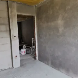 Świeżo otynkowane, szare ściany wewnętrzne w surowym stanie, widoczny otwór drzwiowy z wiadrami i drabiną w tle.