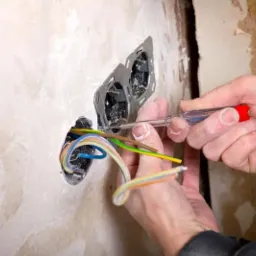 Elektryk sprawdza napięcie w gniazdku za pomocą próbnika napięcia, widoczne kolorowe przewody i surowa ściana z niedokończonym wykończeniem.
