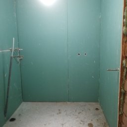 Łukasz Matuszak - Wnętrze łazienki w trakcie prac, ściany wyłożone zielonymi płytami kartonowo-gipsowymi, widoczne otwory na instalacje i elementy mocujące, surowy beton na podłodze z plamami, ściana z cegieł...