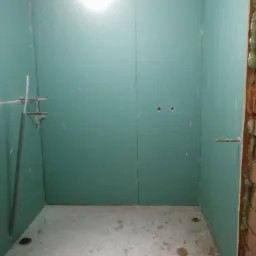 Wnętrze łazienki w trakcie prac, ściany wyłożone zielonymi płytami kartonowo-gipsowymi, widoczne otwory na instalacje i elementy mocujące, surowy beton na podłodze z plamami, ściana z cegieł...
