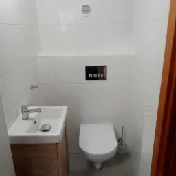 Nowoczesna, mała łazienka z białymi płytkami o falującej strukturze, podwieszaną toaletą, umywalką z szafką z drewnopodobnego materiału i szarą podłogą.