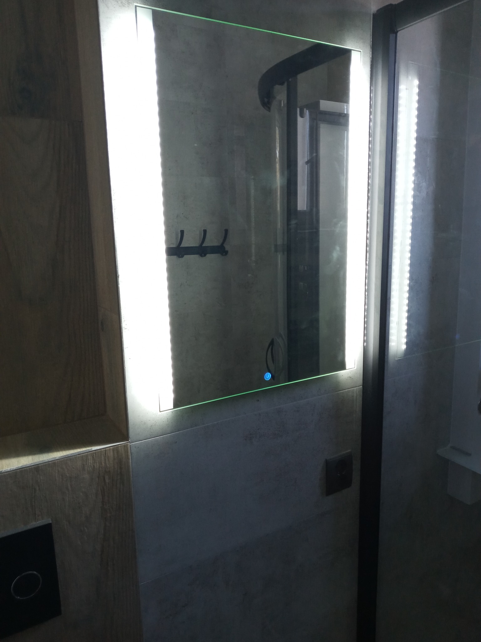 Oświetlone lustro w łazience z szarymi płytkami, widoczne wieszaki na ręczniki i panel sterowania oświetleniem.