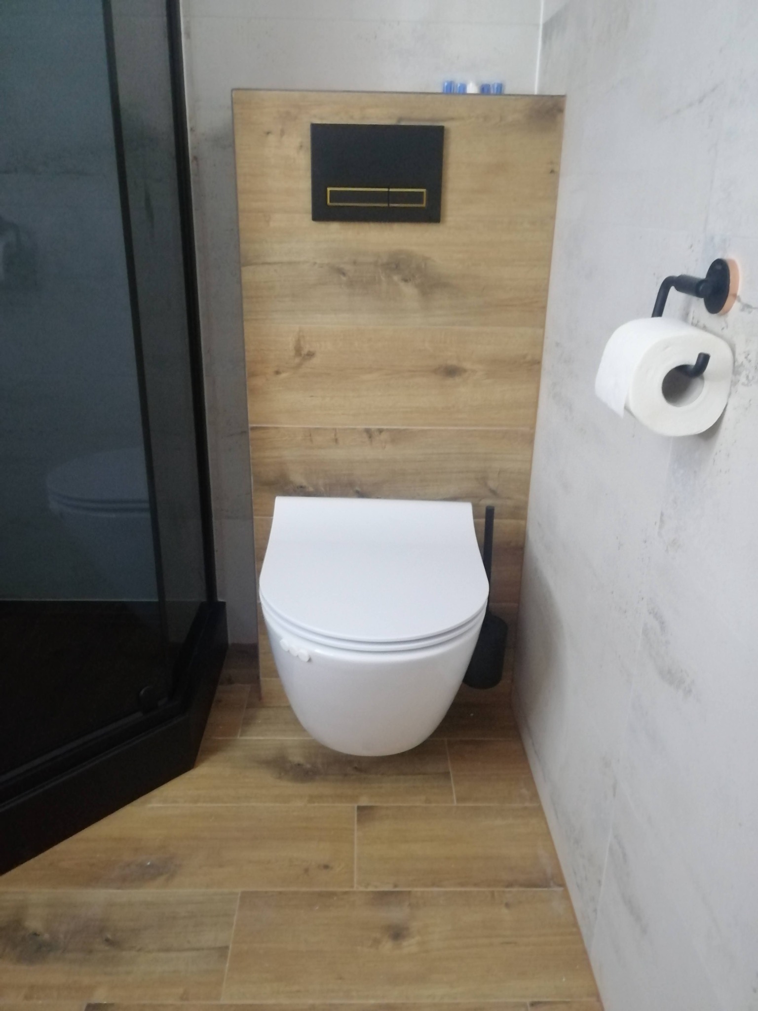 Nowoczesna toaleta podwieszana z czarnym panelem spłukującym na tle drewnopodobnej okładziny, widoczna w łazience w Pleszewie po remoncie.