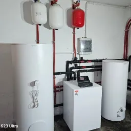 Kompletna instalacja grzewcza w kotłowni: biały kocioł gazowy z panelem sterowania, dwa białe i jeden czerwony zbiornik ciśnieniowy, rozbudowany system rur.