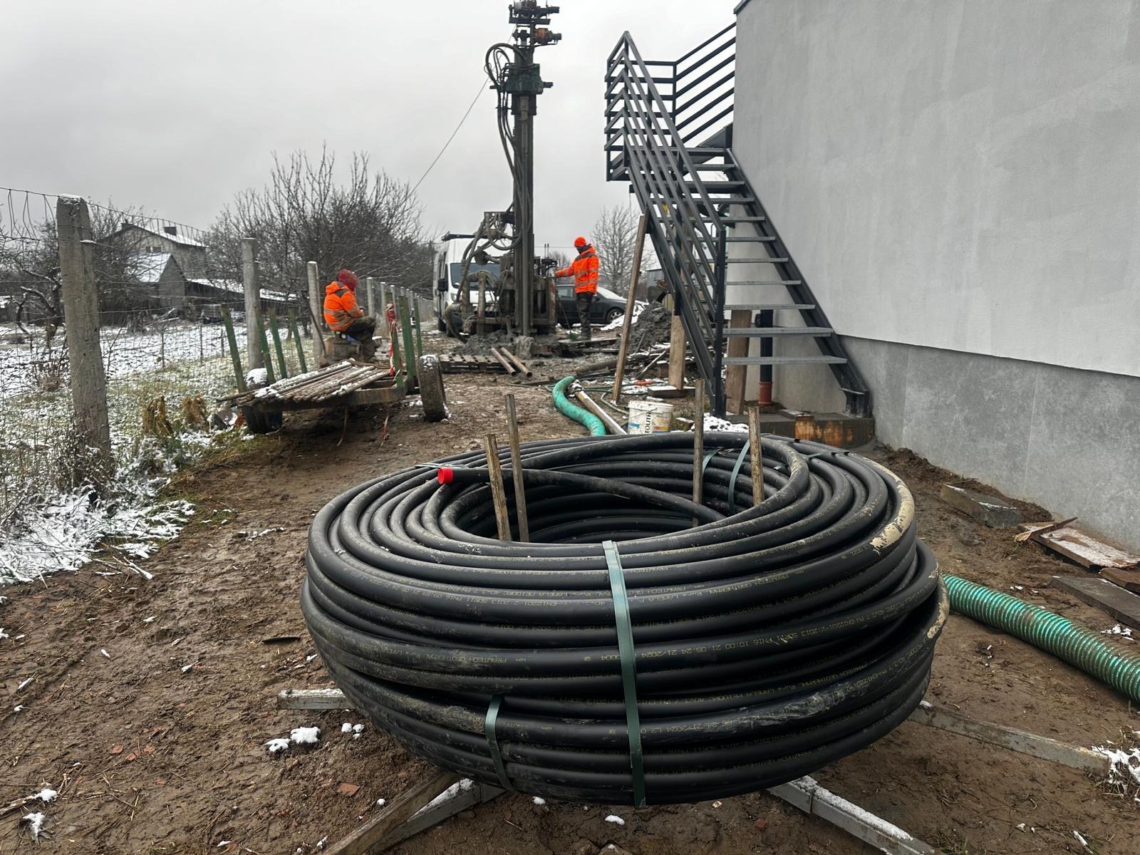 Ziemny wymiennik ciepła w postaci zwiniętej rury na placu budowy, w tle wiertnica i pracownicy w pomarańczowych ubraniach, pochmurny dzień.