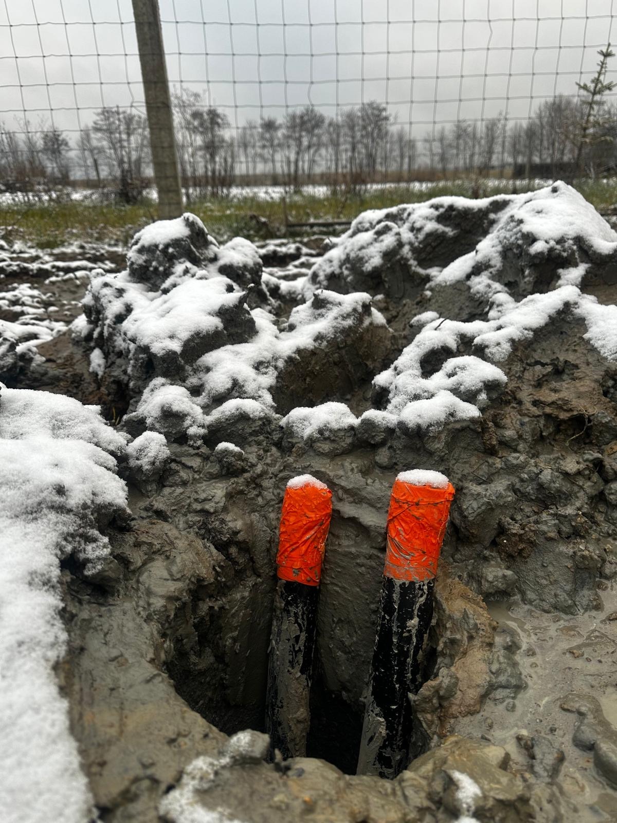 Dwa czarne rury gazowe z pomarańczowymi zaślepkami wystające z wykopu w ziemi pokrytej śniegiem, widoczne ogrodzenie i drzewa w tle.