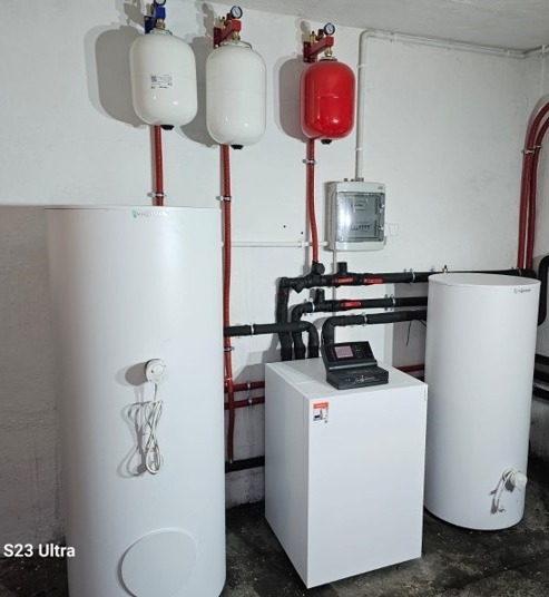 Kompletna instalacja grzewcza w kotłowni: biały kocioł gazowy z panelem sterowania, dwa białe i jeden czerwony zbiornik ciśnieniowy, rozbudowany system rur.