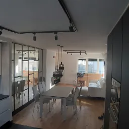 Nowoczesne wnętrze mieszkania z otwartą kuchnią, jadalnią i salonem, widoczne białe szafki kuchenne z czarnym blatem, szklana ściana działowa z czarnymi szprosami, stół z krzesłami, biała sofa...