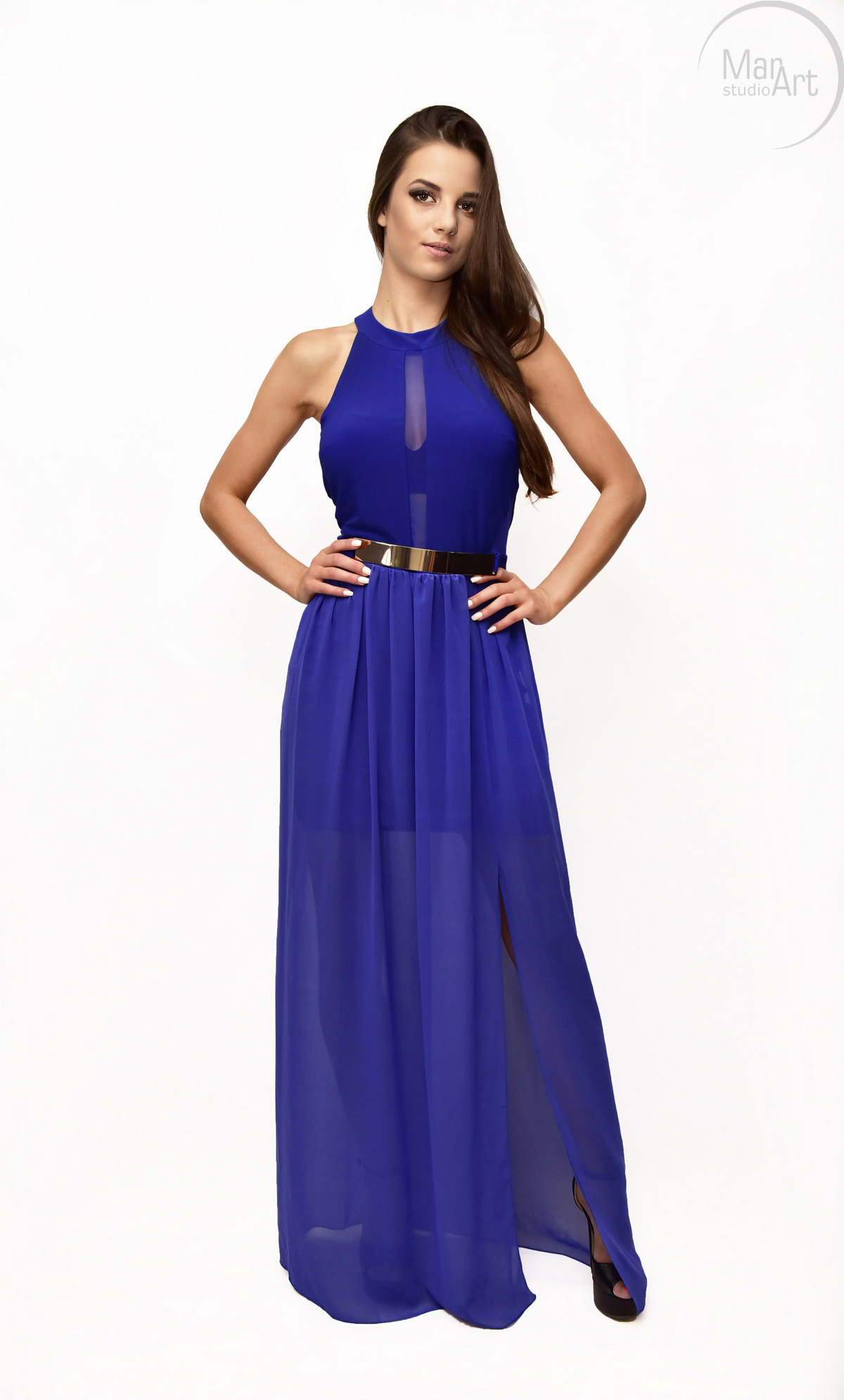 Kobieta w długiej, kobaltowej sukni z wysokim rozcięciem i złotym paskiem, pozująca na białym tle w studiu.