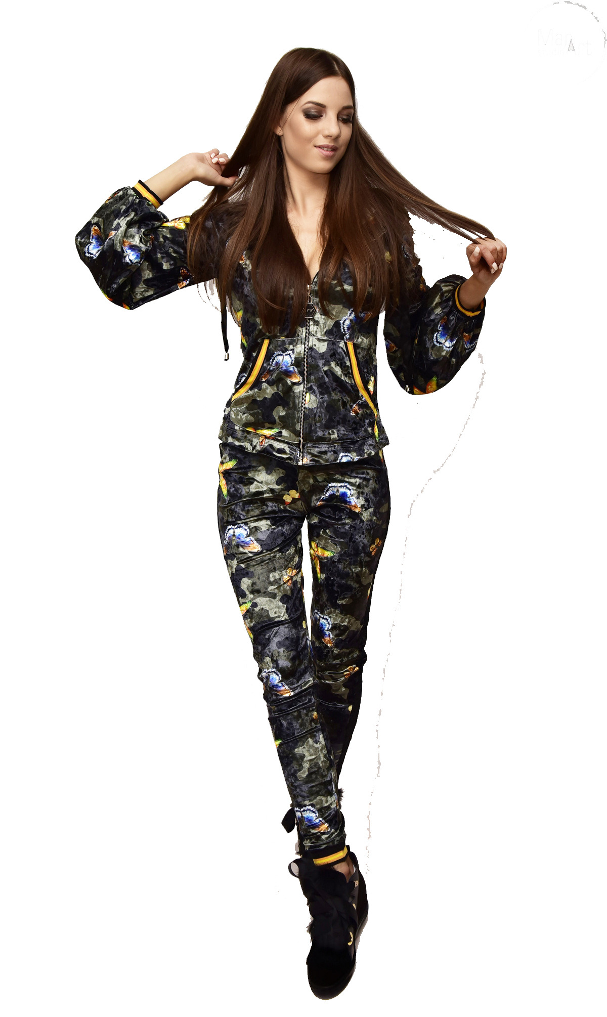 Kobieta pozuje w dresie z motywem kamuflażu i motyli, na białym tle, z delikatnym uśmiechem i dłonią we włosach.