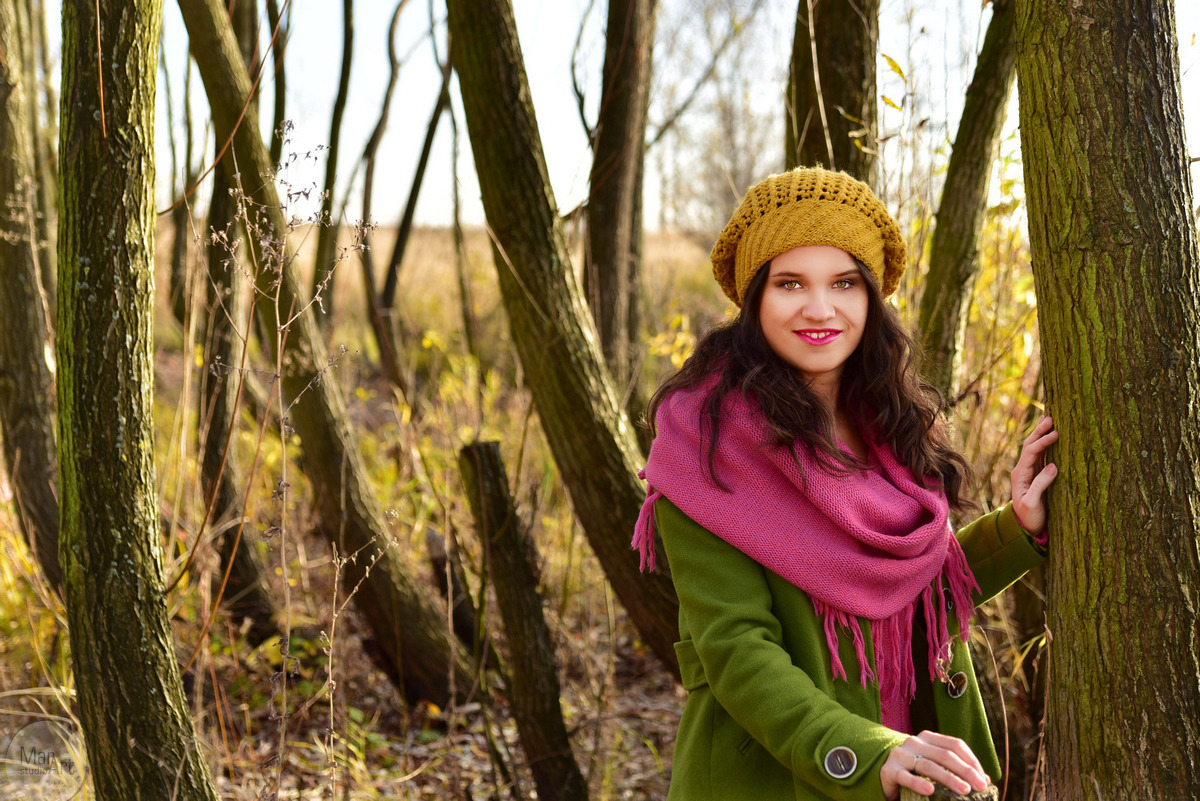 Portret kobiety w jesiennej scenerii, oparta o drzewo, w zielonym płaszczu, różowym szalu i żółtej czapce, z delikatnym uśmiechem i makijażem.