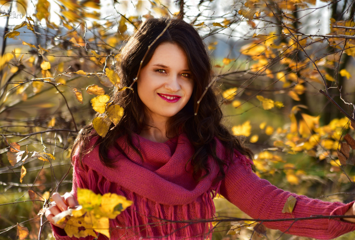 Portret uśmiechniętej kobiety w różowym swetrze z frędzlami, otoczonej gałązkami z żółtymi liśćmi w jesiennym słońcu.