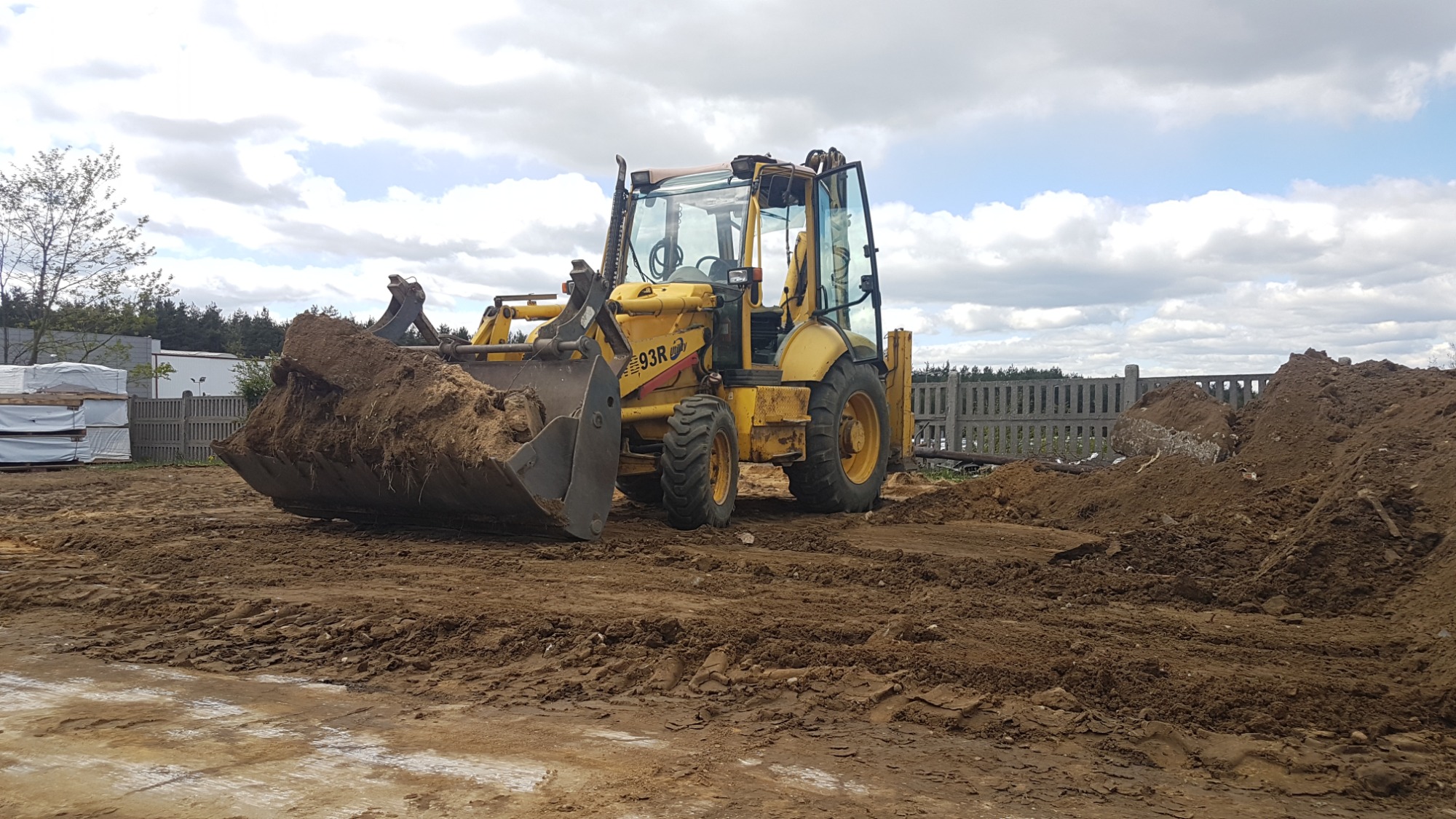 Żółta koparko-ładowarka z pełną łyżką ziemi na placu budowy, widoczne hałdy ziemi i ogrodzenie w tle.