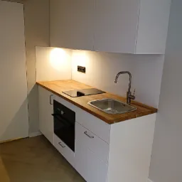 Minimalistyczna zabudowa kuchenna z białymi szafkami, drewnianym blatem, stalowym zlewem i czarną płytą indukcyjną, widoczne gniazdka elektryczne i oświetlenie podszafkowe.