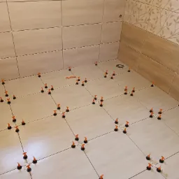 Układanie jasnych, beżowych płytek podłogowych i ściennych z użyciem pomarańczowych klin&oacute;w poziomujących w narożniku łazienki z odpływem podłogowym.