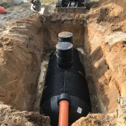 Czarny zbiornik z dwoma włazami w wykopie, podłączony pomarańczową rurą, widoczny minikoparka Bobcat E16 i betonowy słup w tle.