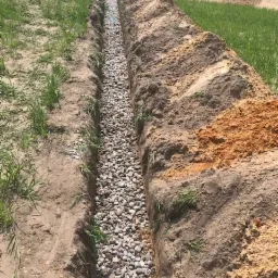 Wykop wypełniony żwirem, prawdopodobnie w celu odprowadzenia wody, widoczne rury drenażowe w oddali, świeżo usypana ziemia obok rowu.