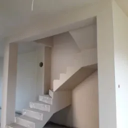 Wnętrze domu w trakcie prac wykończeniowych: surowe schody betonowe, ściany po nałożeniu gładzi, widoczny kabel zwisający z sufitu.